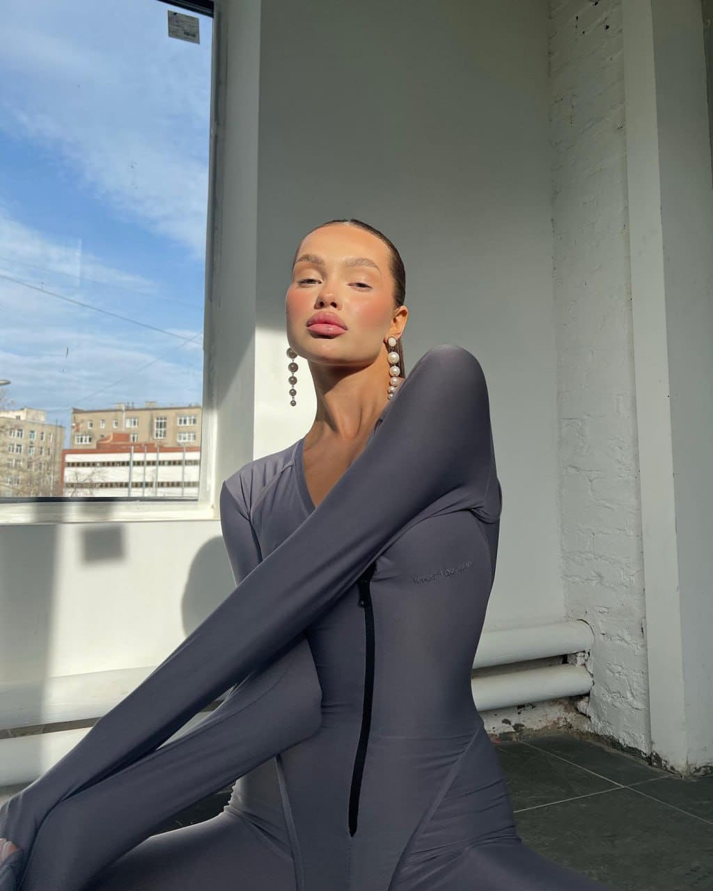 A woman with slicked-back hair sits in a sunlit room wearing a grey bodysuit and dangling earrings, arms crossed.