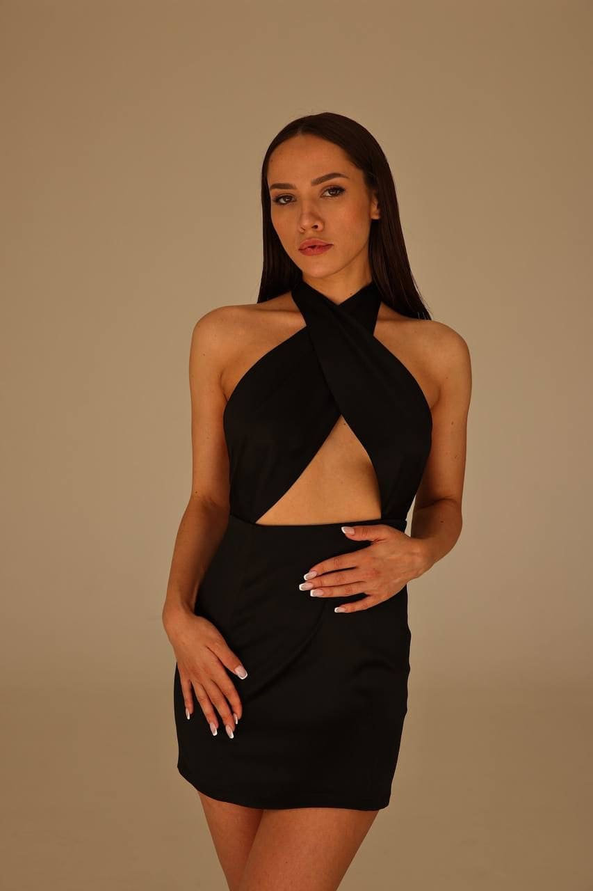 Vera stands against a beige backdrop in a black halter-neck mini dress with a front cutout, hands on hips and thighs.