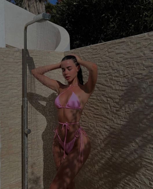 A woman in a pink string bikini poses with hands behind her head outdoors, standing near a shower fixture and a textured stone wall.