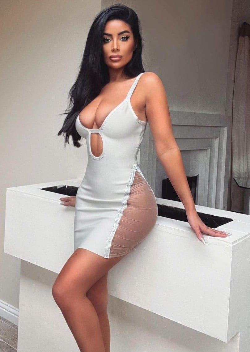 A woman leans against a white counter wearing a short white dress with sheer side panels, with a fireplace visible in the background.