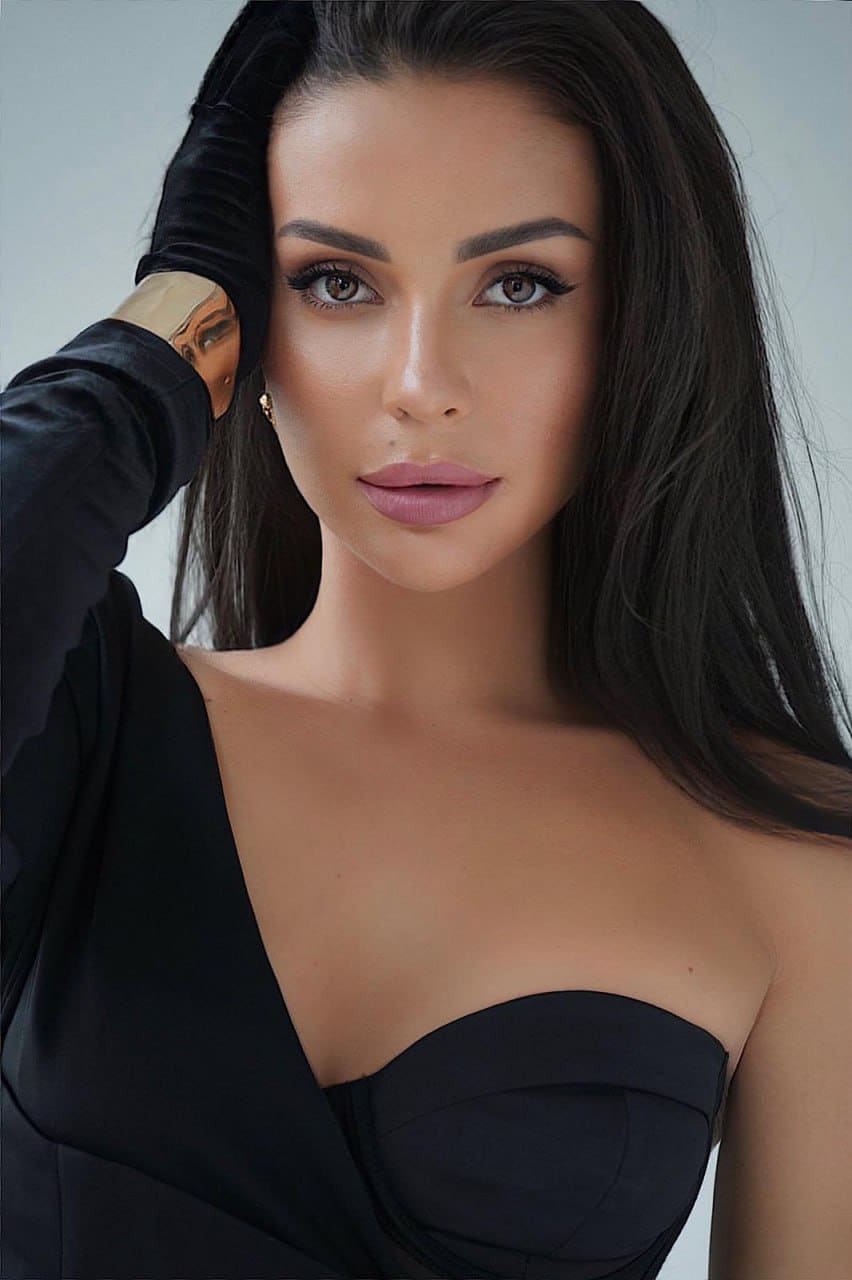 A woman in a black one-shoulder top raises her gloved right hand to her head, wearing a gold cuff, against a plain light background.