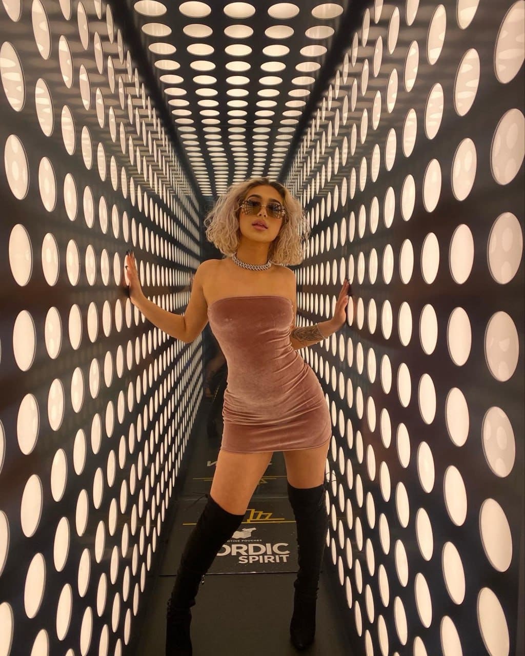 A woman in a mauve velvet dress and black thigh-high boots presses her hands against walls in a lit corridor.