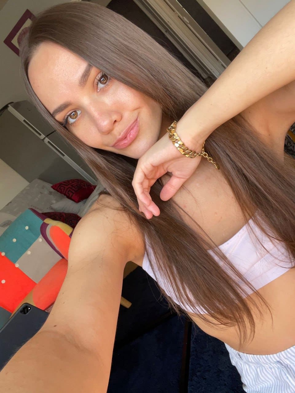 Bedroom selfie wearing a white strapless top and gold bracelet, with one arm raised and the other holding the camera.