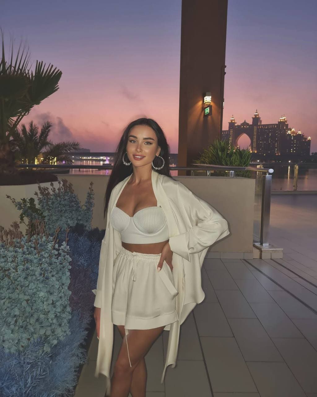 A woman in a white bralette, shorts, and open jacket stands on a tiled terrace at dusk with a city skyline behind her.