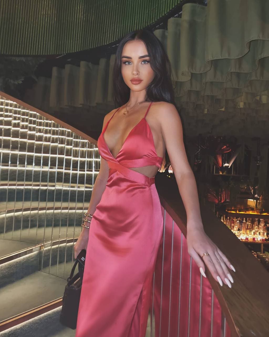 A woman in a deep V-neck pink satin dress with cut-outs stands on an indoor staircase, holding a black handbag and railing.