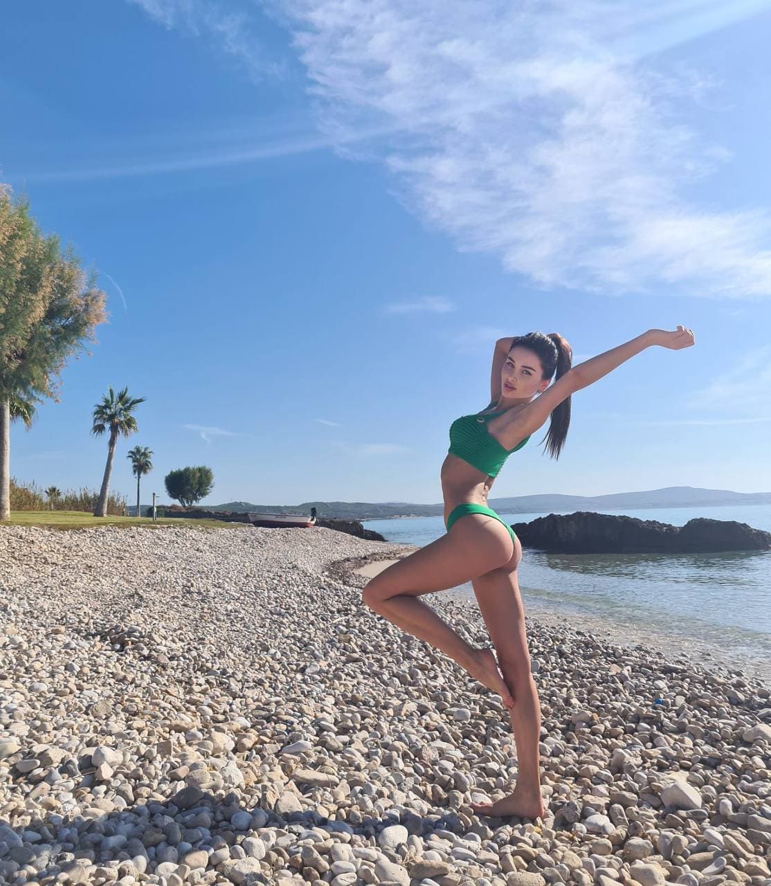 A barefoot woman in a green two-piece swimsuit stretches on a pebble beach with palm trees and calm sea in the background.
