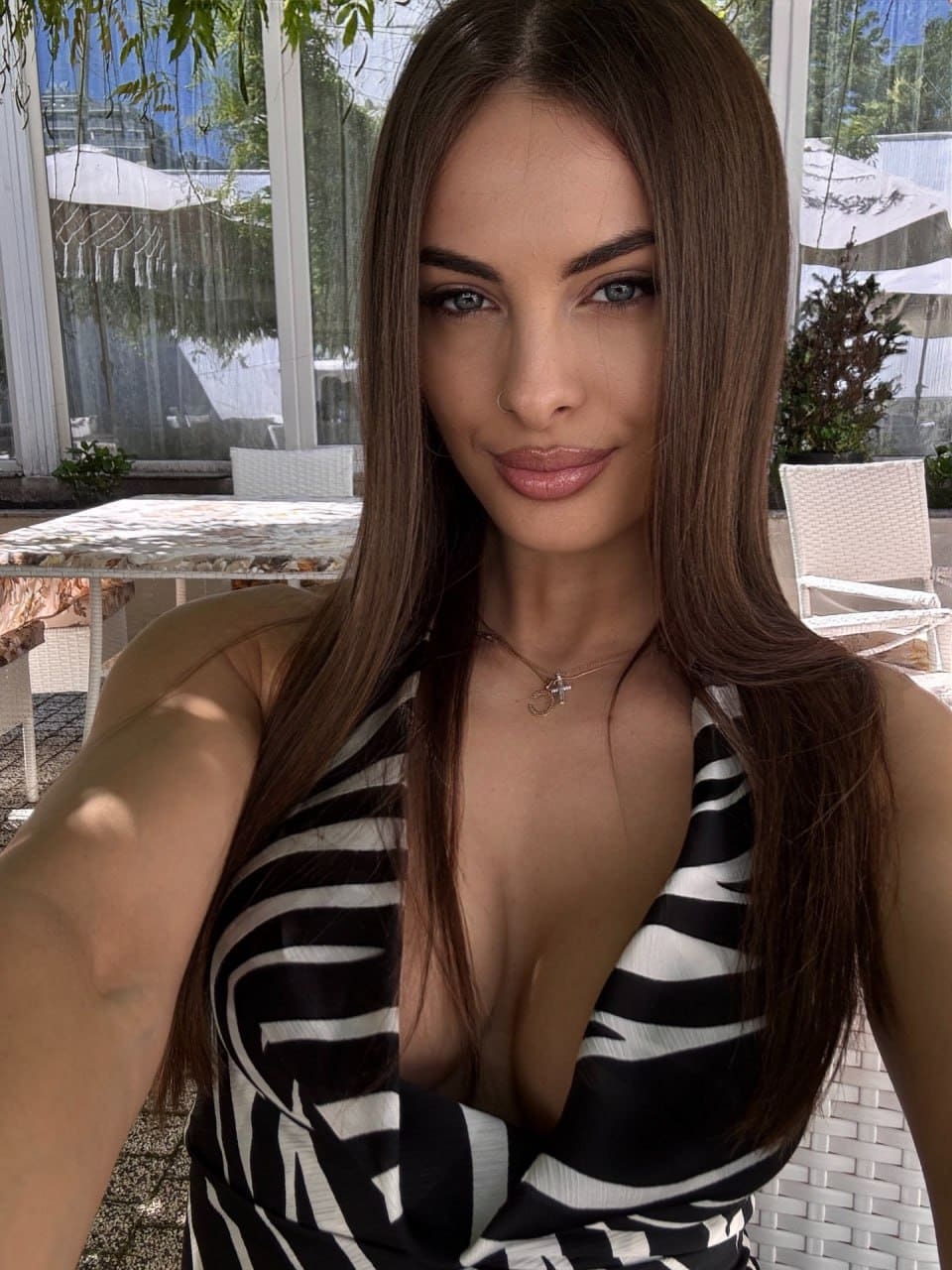 Elizabeth wears a black-and-white patterned halter top with a deep V-neckline, posing outdoors near white wicker furniture and glass windows.