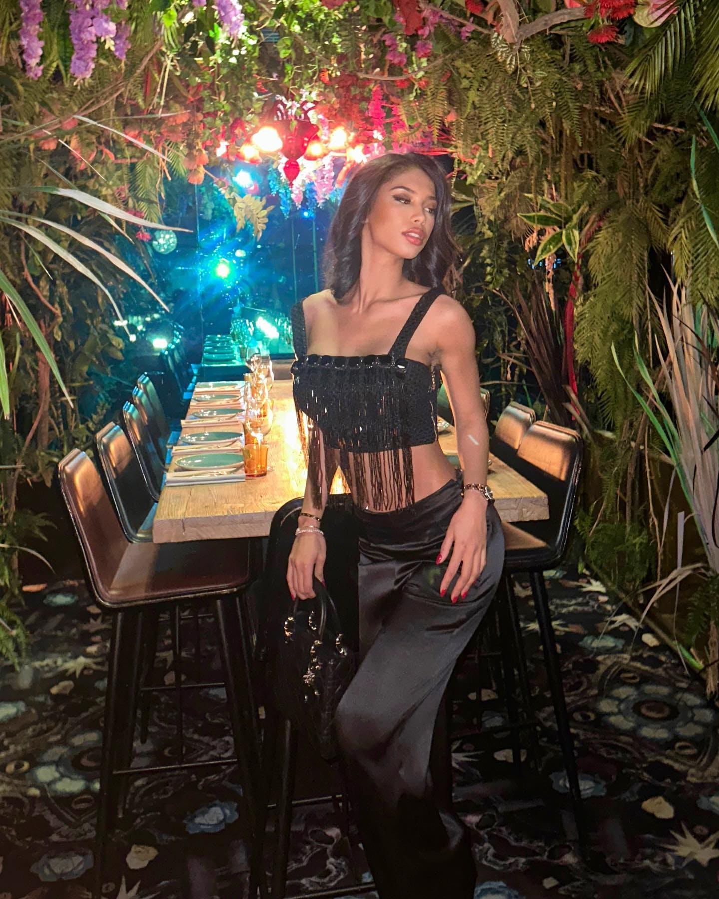A woman sits on a stool in a plant-filled venue, wearing a black fringed crop top and satin trousers while holding a black handbag.