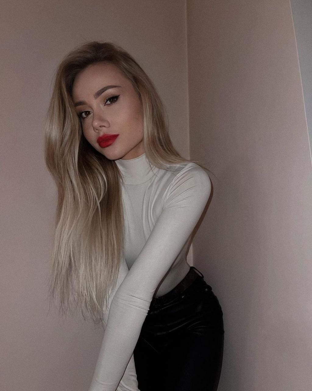 Hazel stands in a corner wearing a white turtleneck and black pants, posing with arms crossed.