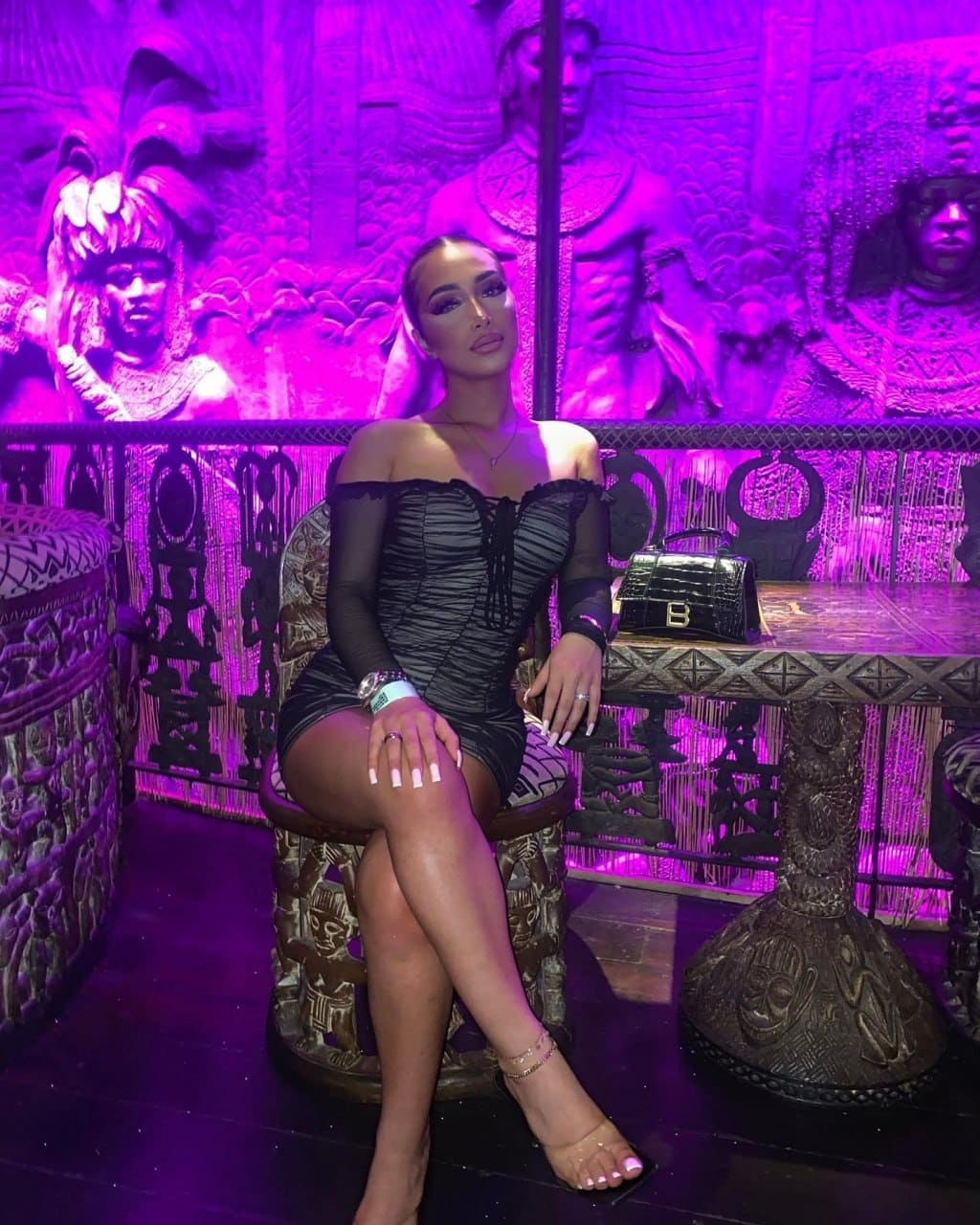 A woman with dark hair sits on an ornate stool in a black dress with sheer sleeves in a purple-lit room.