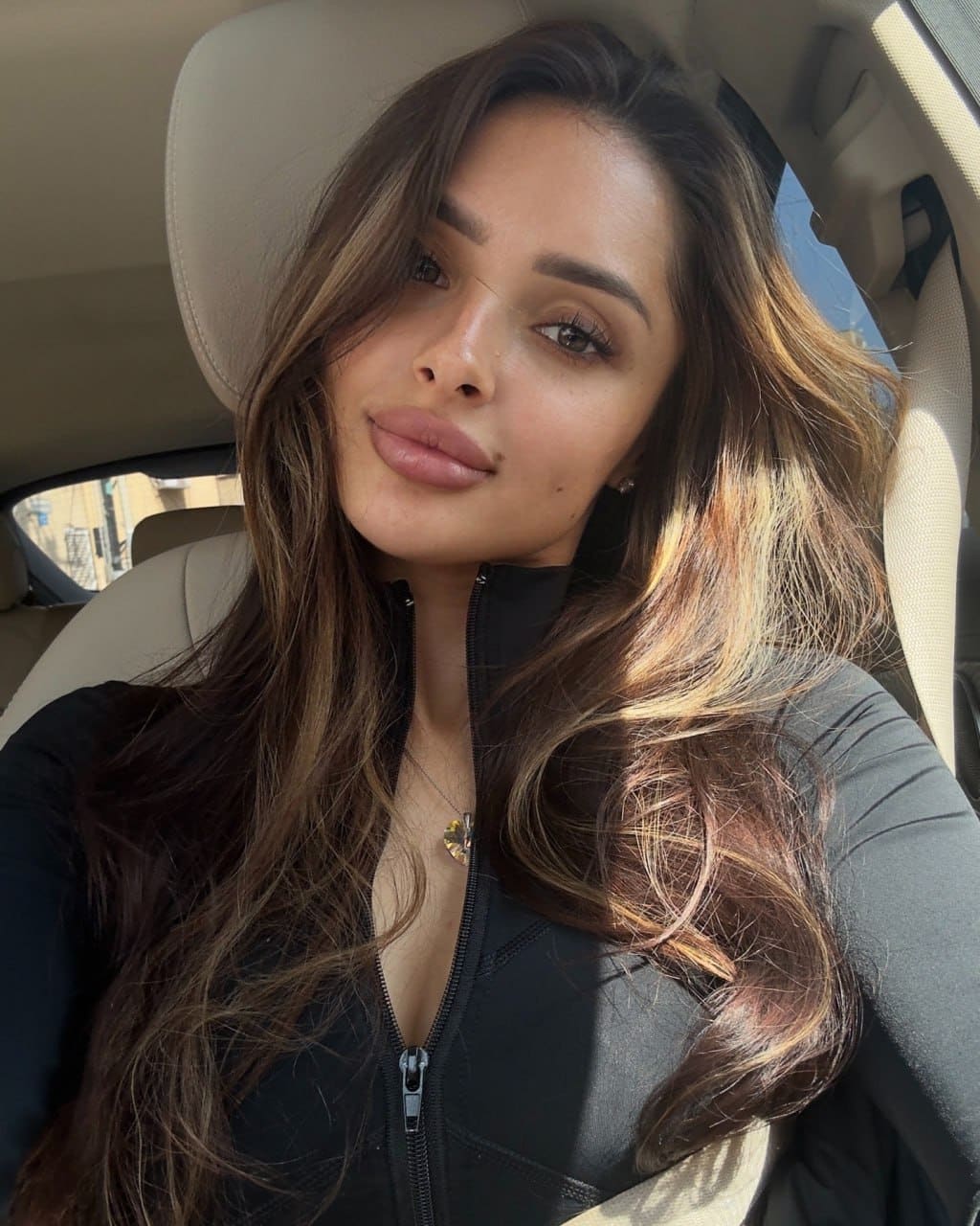 Oli sits in a car driver's seat, wearing a partially unzipped black top and necklace, with sunlight illuminating her face and wavy brown hair.