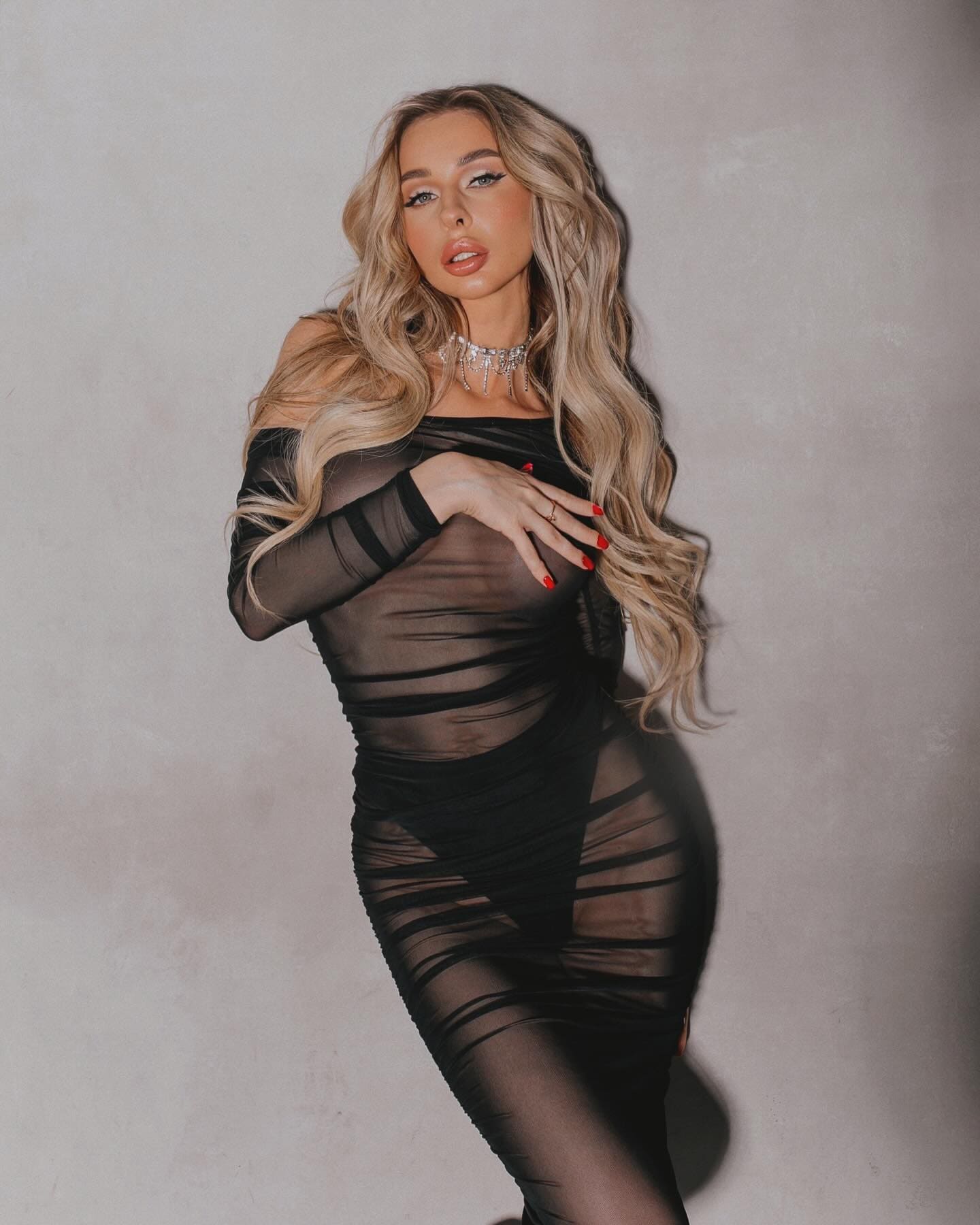 Stands against a gray wall in a sheer black dress with cutouts, hand on chest, wearing a sparkling choker.