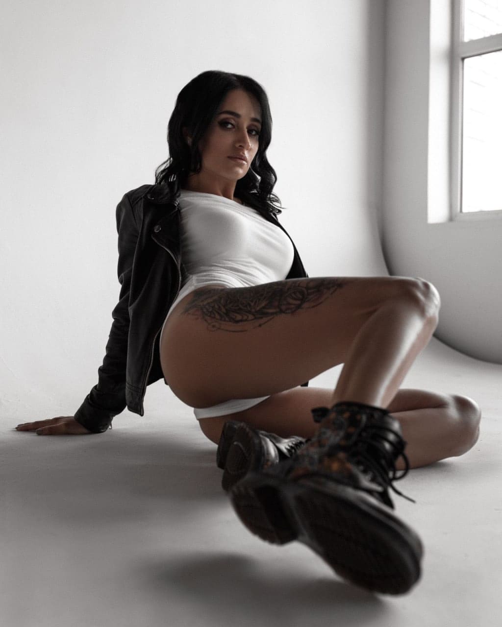 A woman sits on a gray floor in a white room, wearing a white bodysuit, open leather jacket, and boots, showing a floral thigh tattoo.