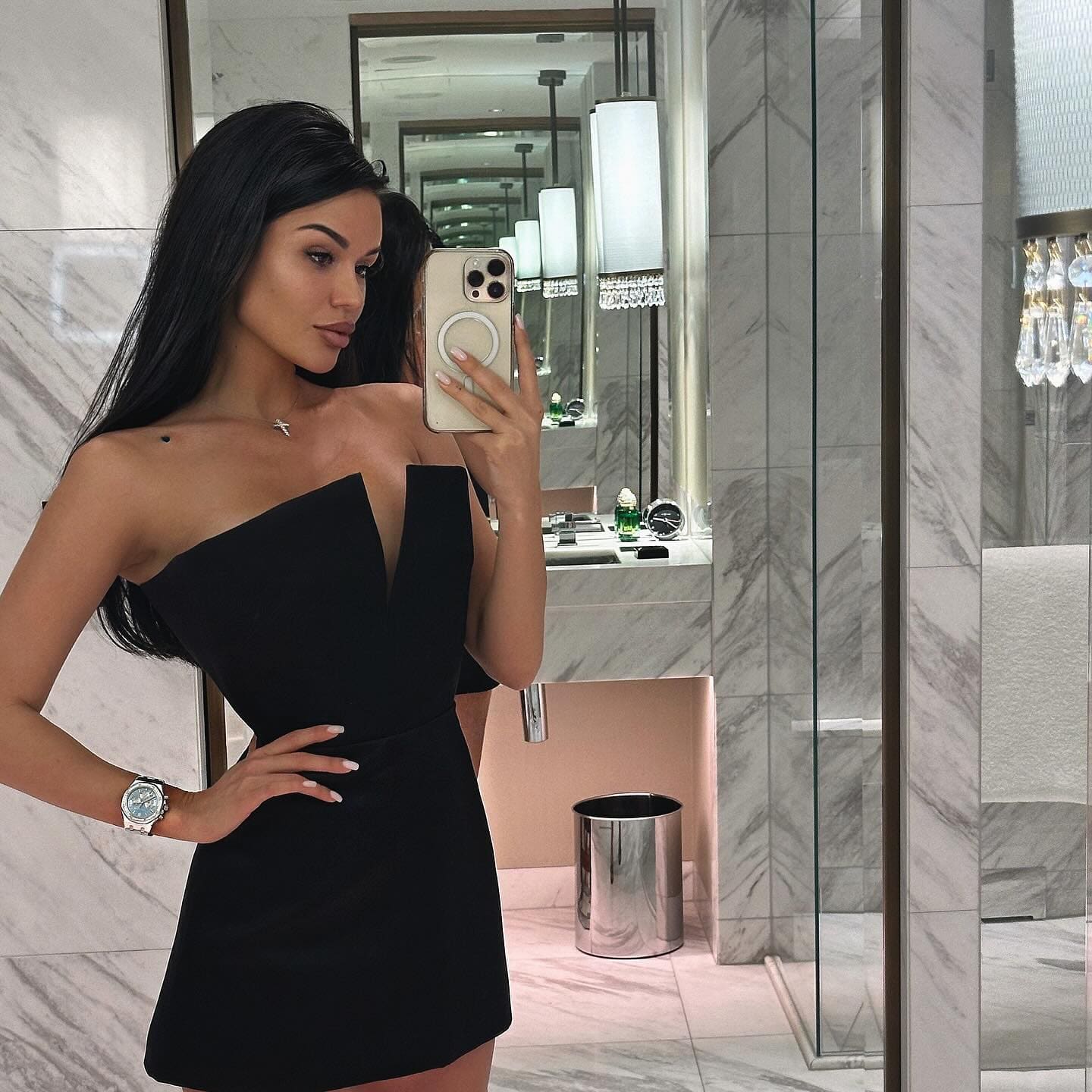 Irma takes a mirror selfie in a marble bathroom, wearing a short black strapless dress with a deep V-neckline.