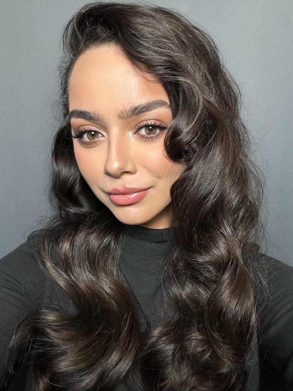 Adama with long dark wavy hair in a black high-neck top smiles slightly against a plain gray background.