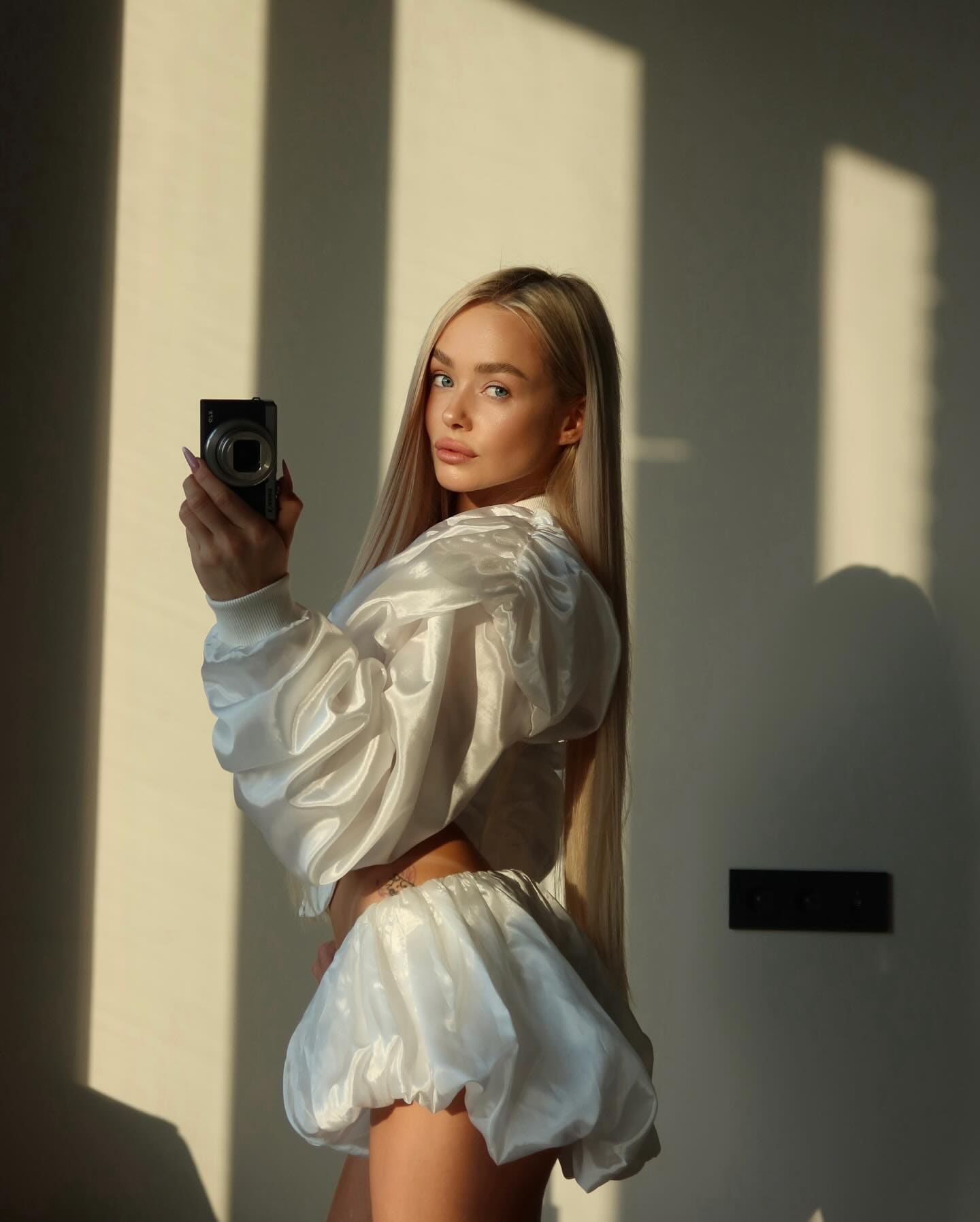 A blonde woman in a white satin crop top and shorts holds a black camera while standing in a sunlit room.