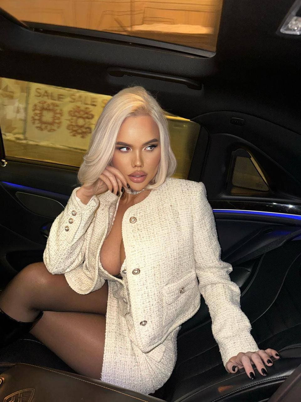 A blonde woman in a white tweed jacket and stockings sits in a car back seat, one hand near her face.