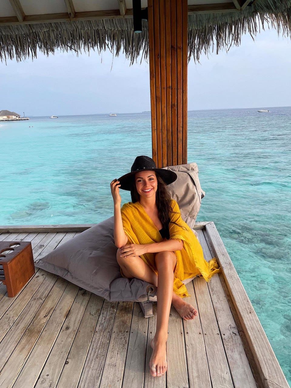 A woman in a yellow wrap dress and black hat sits cross-legged on a deck, touching her hat brim against a backdrop of ocean water.