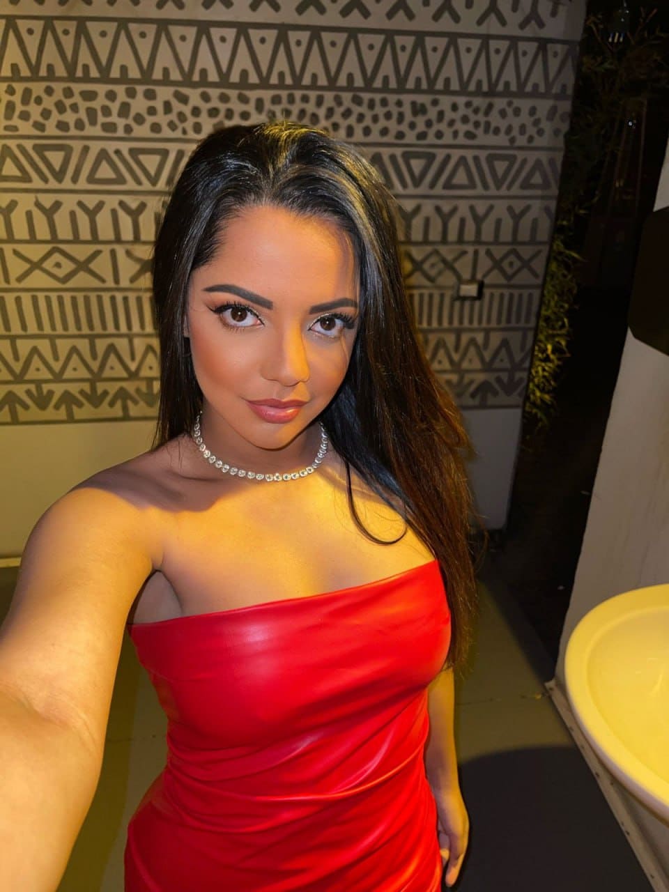 A woman takes a selfie in a red strapless dress and beaded necklace, standing before a wall with a geometric pattern.