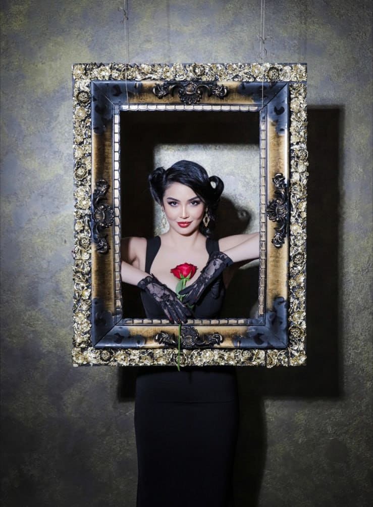 A woman with side buns and lace gloves stands behind a gold frame, holding a red rose against a gray wall.