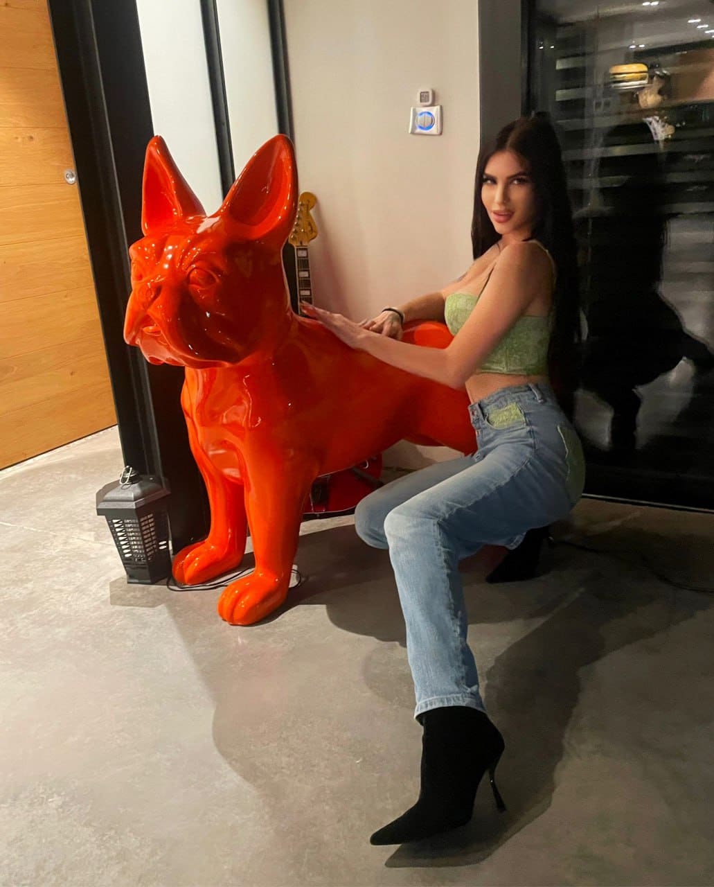 Woman sits on an orange dog sculpture indoors, wearing a green cropped tank top, jeans, and black heeled boots near a glass wall.