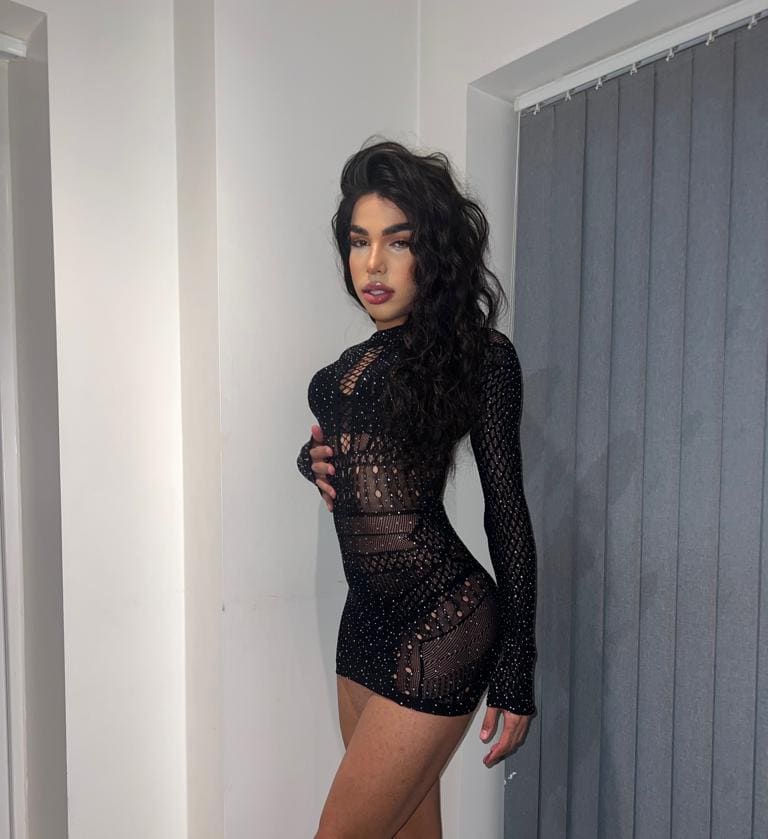A woman stands in a room with white walls and a gray curtain, wearing a long-sleeved black mesh mini dress with cutouts, her hand resting on her chest.