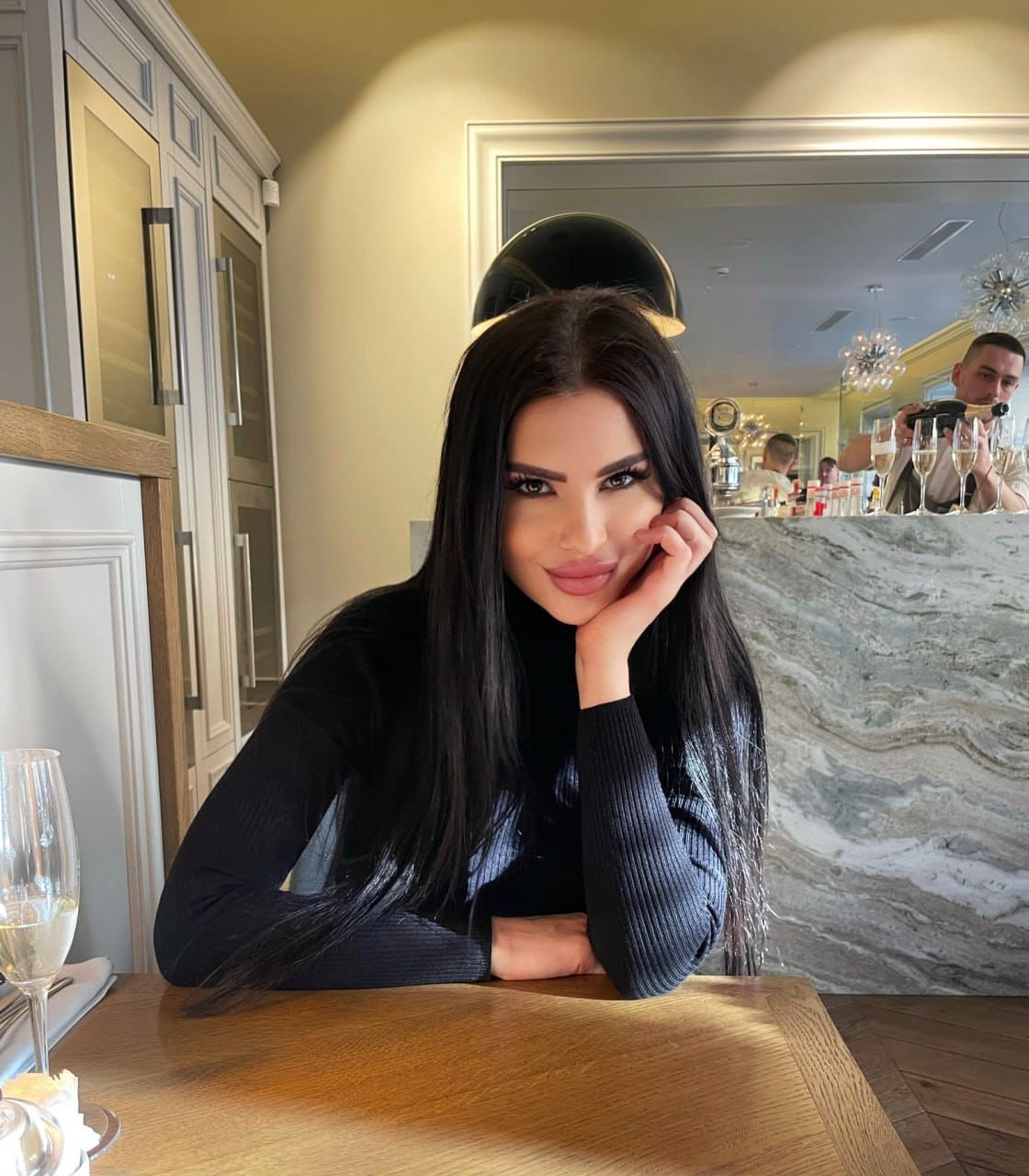 Zoi sits at a restaurant table in a black long-sleeved top, resting her chin on her hand with a wine glass in the foreground.